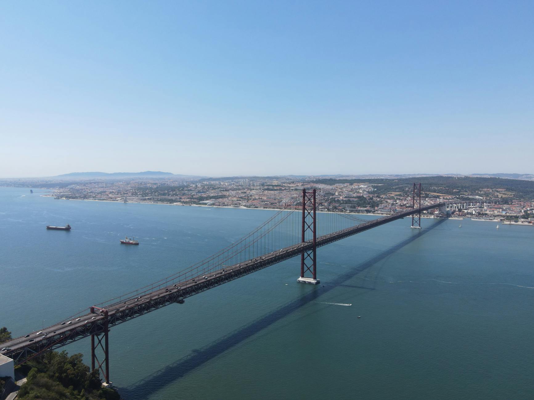 A stunning aerial view of the 25 de Abril Bridge connecting Lisbon across the Tagus River, under a clear blue sky.