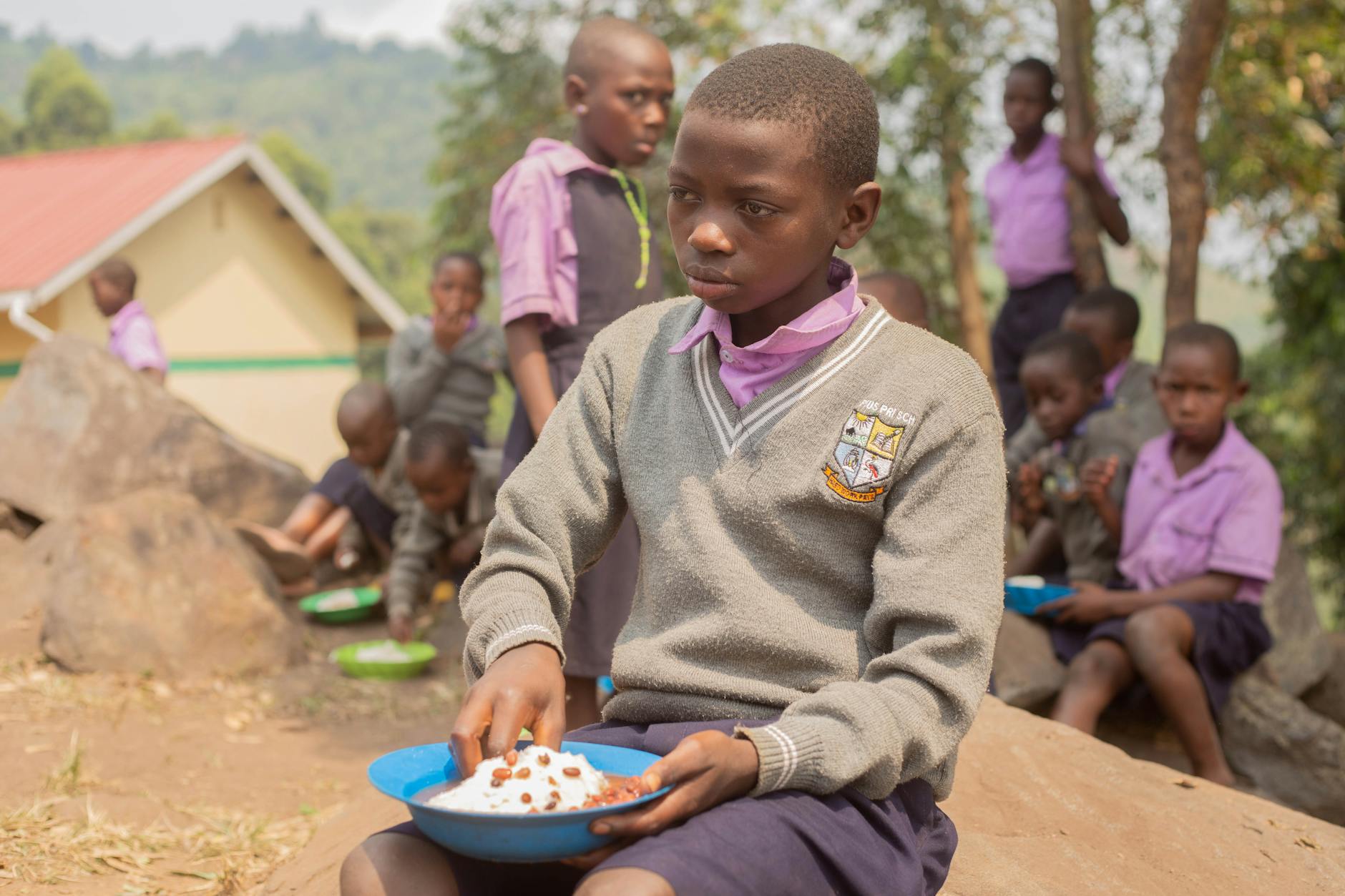 African school children enjoy a meal outdoors in a rural setting, wearing uniforms and sitting on rocks.