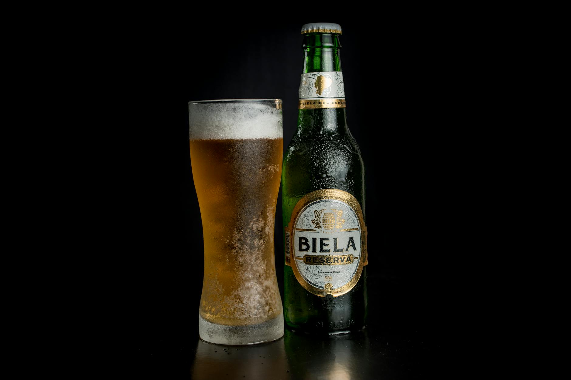 Cold Biela Reserva beer bottle and glass with foam on a black background.