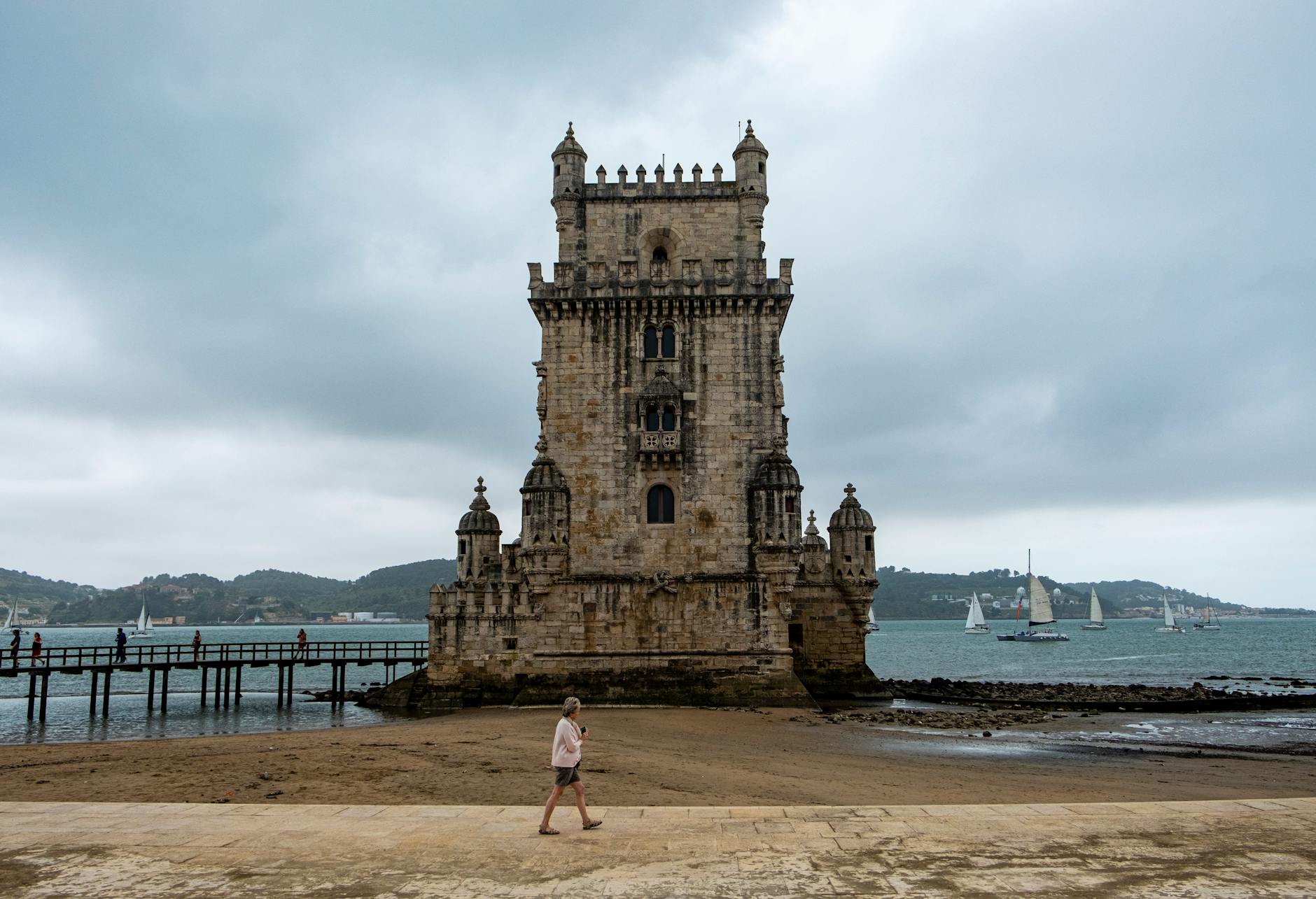 A cloudy day at Torre de Belém, Lisbon, with a scenic seaside view and a walking woman.