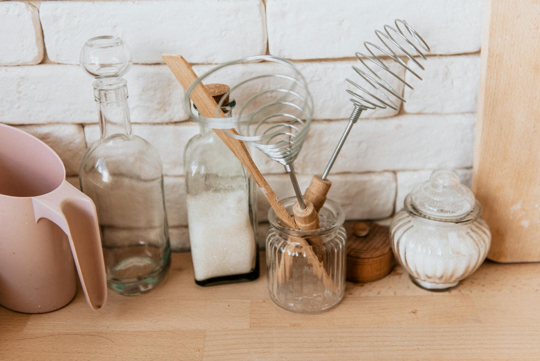 A collection of glass jars and kitchen utensils on a rustic wooden counter with a white brick background.