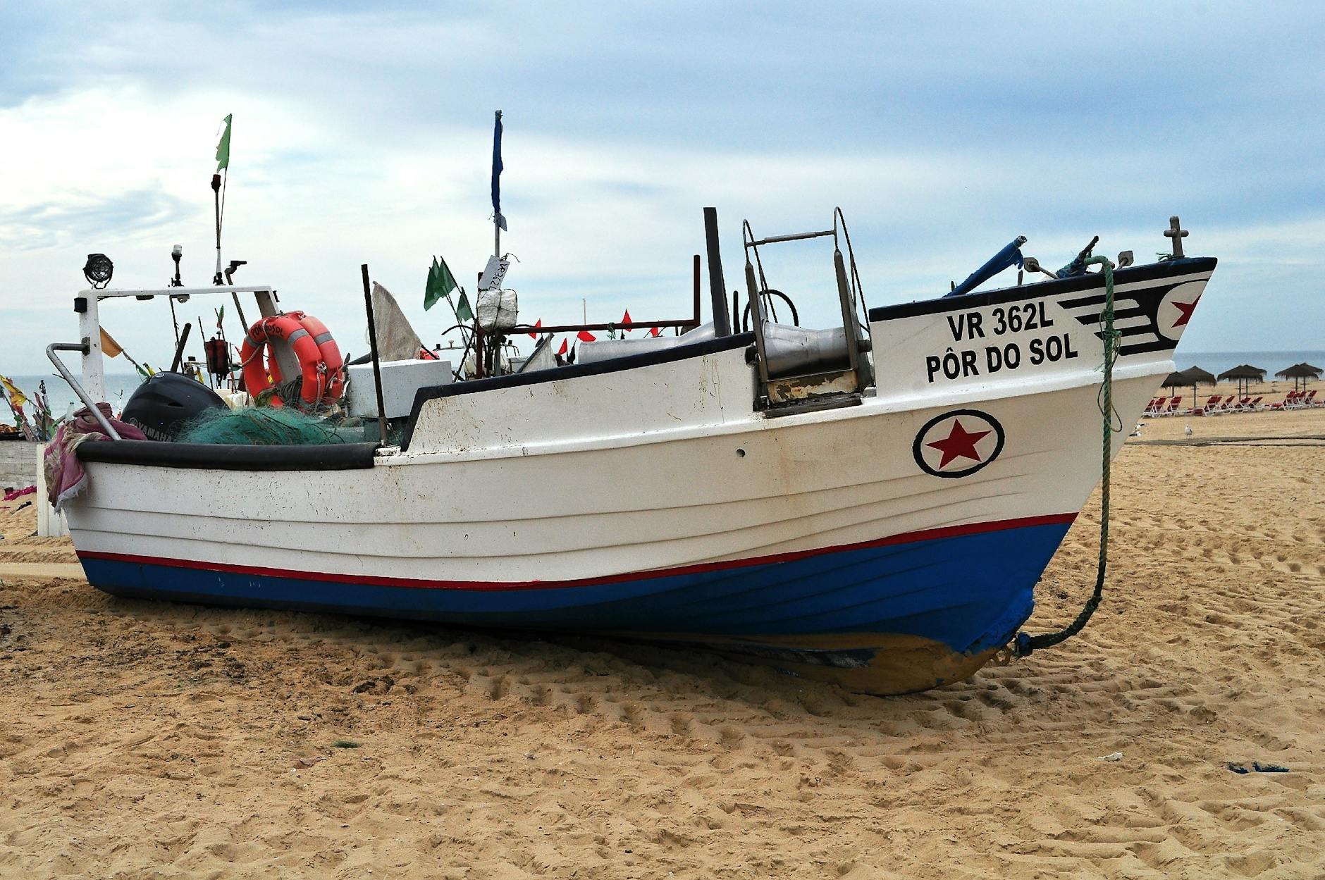 Pôr do sol em Lisboa sem pagar: os miradouros que nunca falham 6 A fishing boat named Pôr do Sol on a sandy beach under a cloudy sky, featuring fishing equipment and nearby umbrellas.