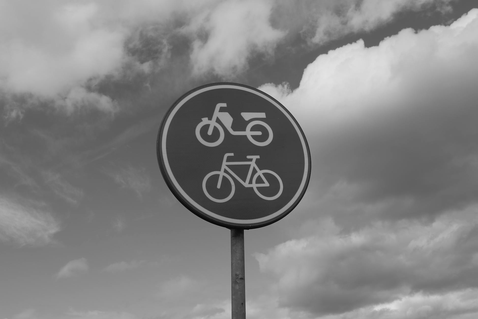 A high-contrast image of a circular road sign indicating bicycle and motorcycle passage, set against a dramatic cloudy sky.