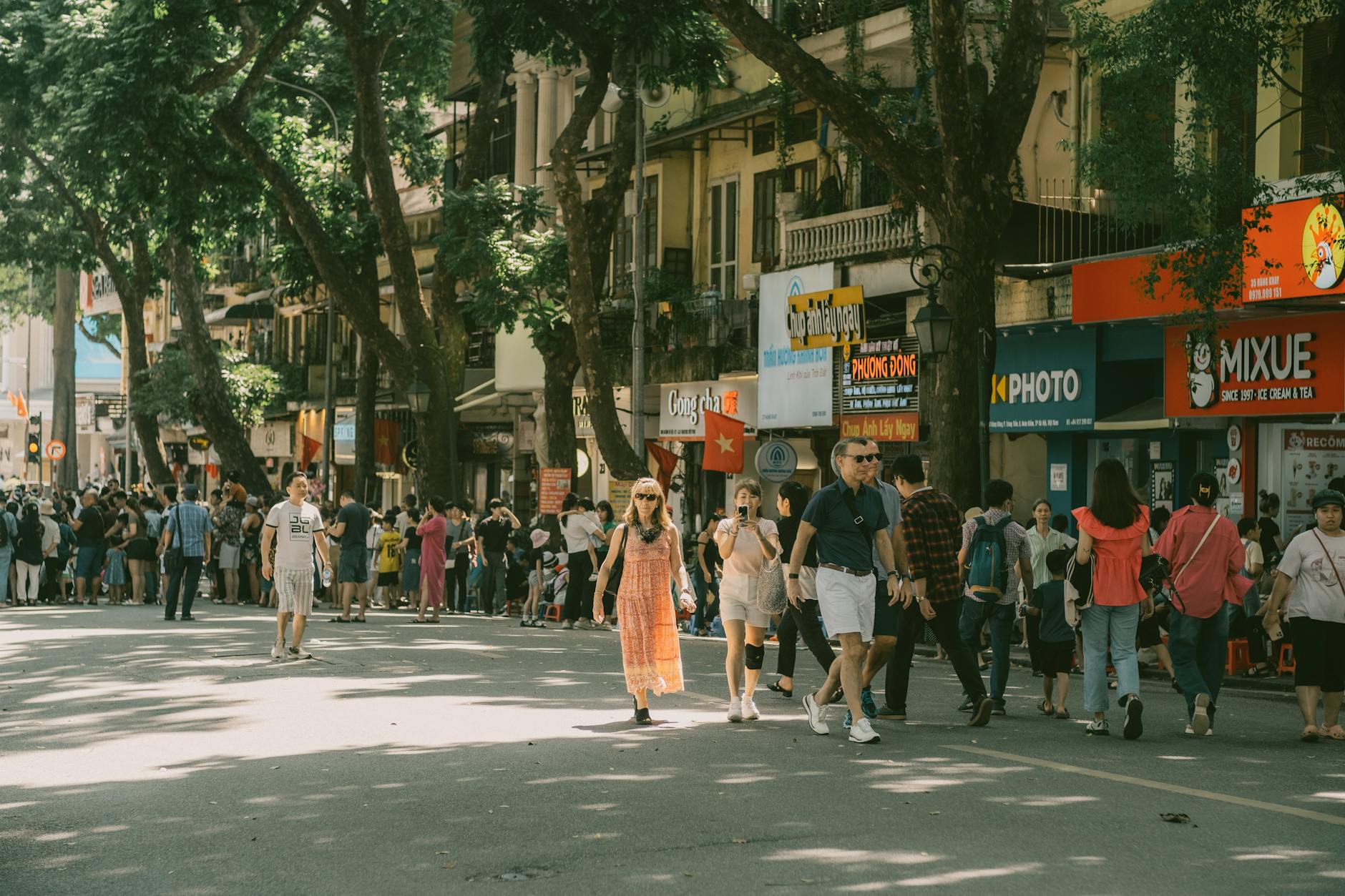 A lively city street filled with people walking under the shade of trees in an Asian city.