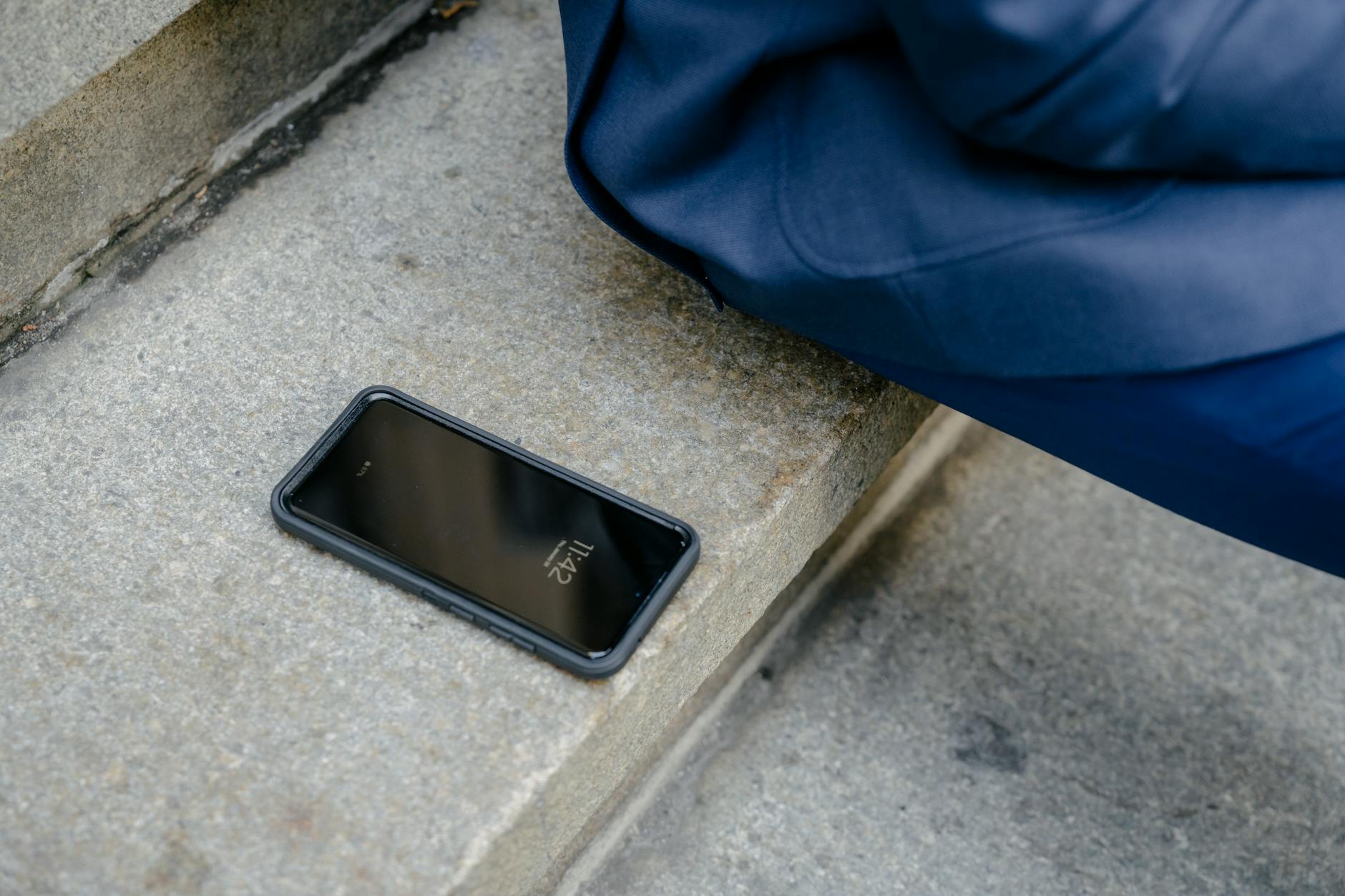 A smartphone is left unattended on concrete steps outdoors, next to a person in blue clothing.