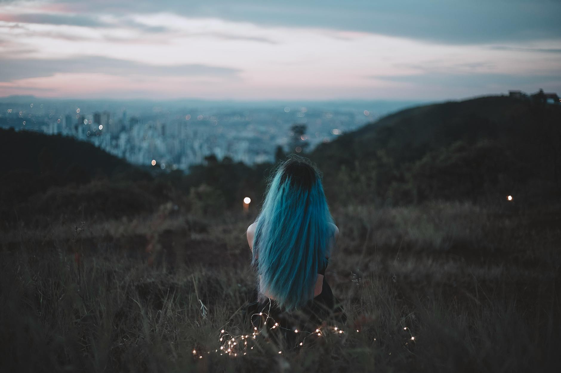 Operafest: como garantir uma noite memorável nos jardins do Palácio do Marquês de Pombal 5 A woman with blue hair sits on a hillside, gazing at the city's twilight skyline adorned with string lights.
