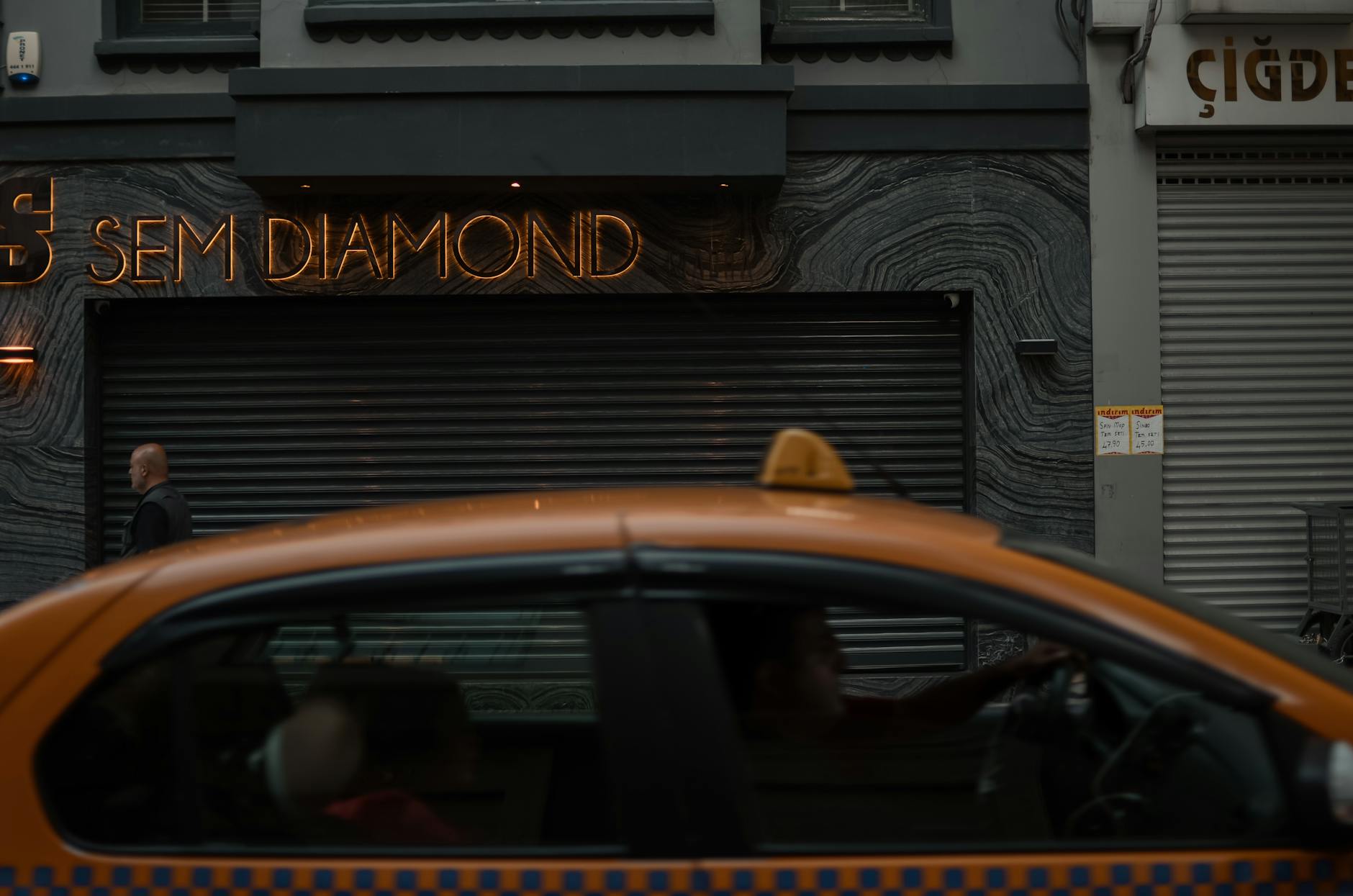 A yellow taxi passing a storefront with neon signage 'SEM DIAMOND' in an urban setting, capturing city life.