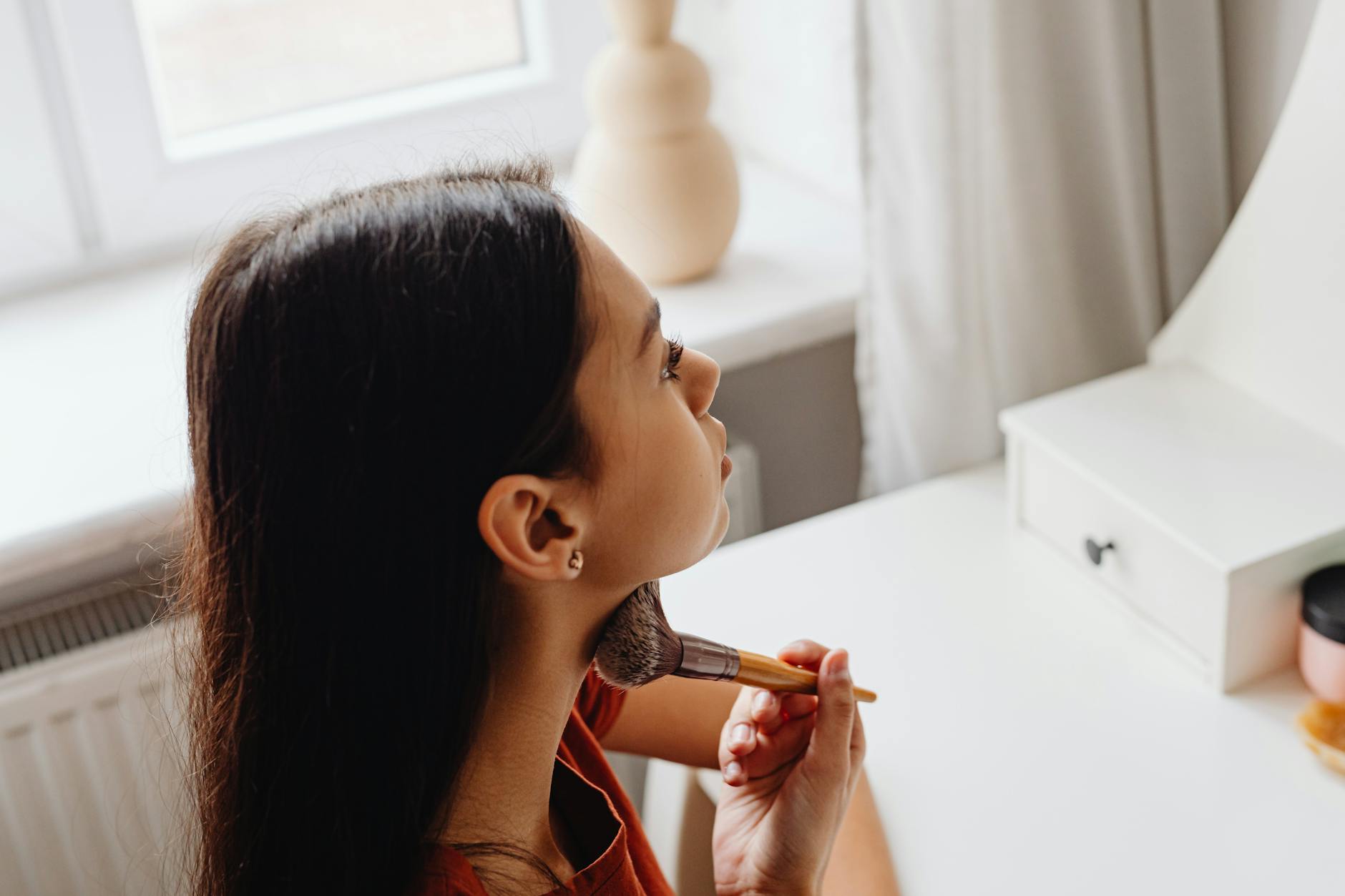 A young woman applies makeup with a brush by her vanity in a warm, indoor setting.