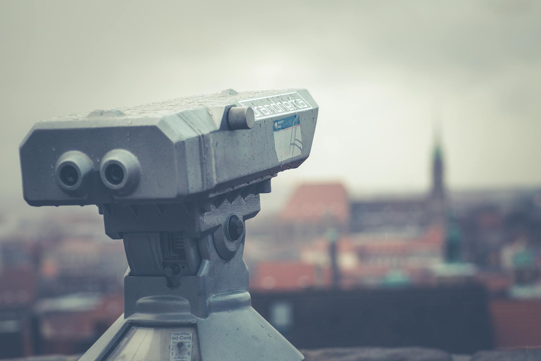 An outdoor telescope overlooking a blurred urban cityscape under a cloudy sky.