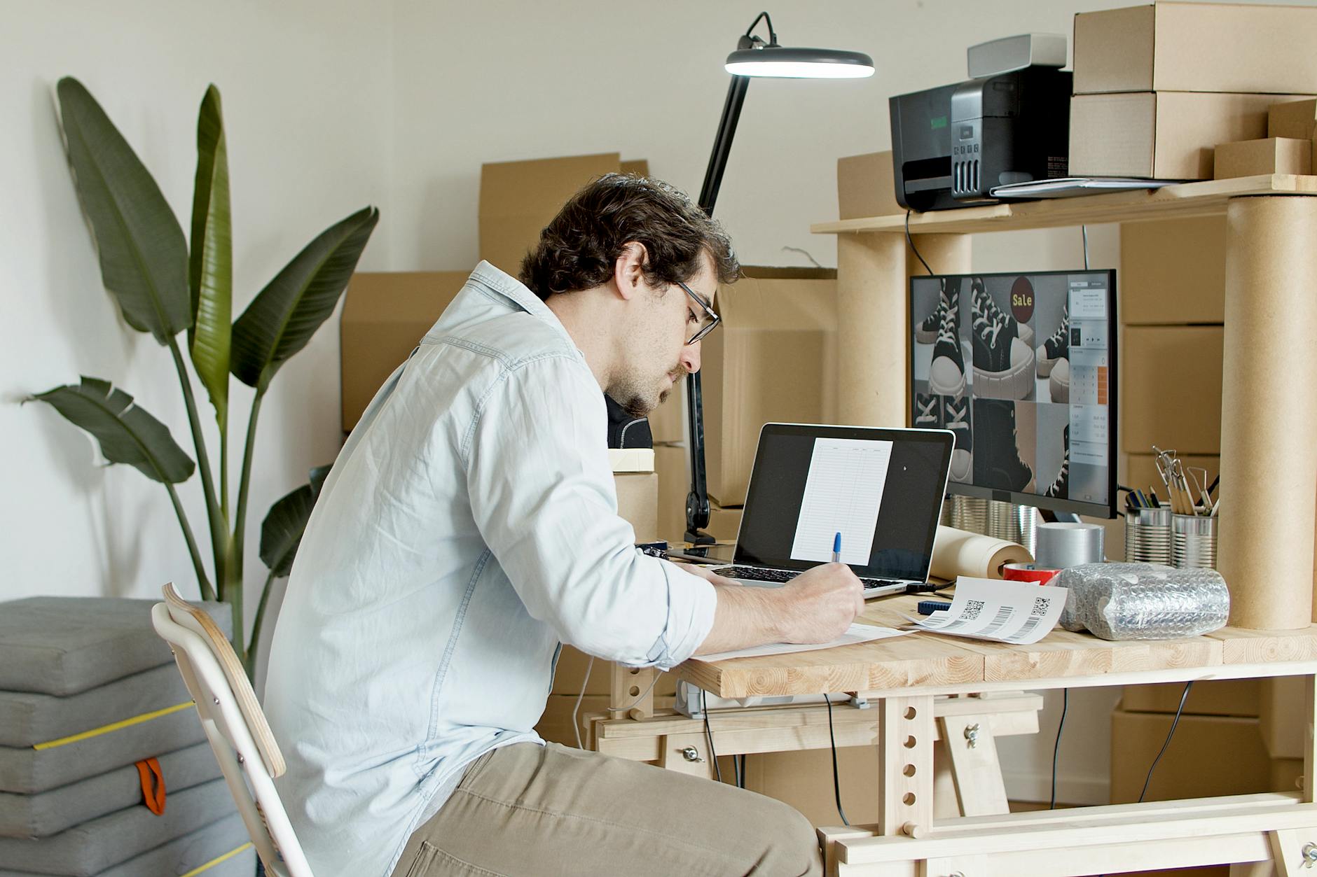 Businessman managing an online store in a home office setting, typing on a laptop with packaging supplies around.