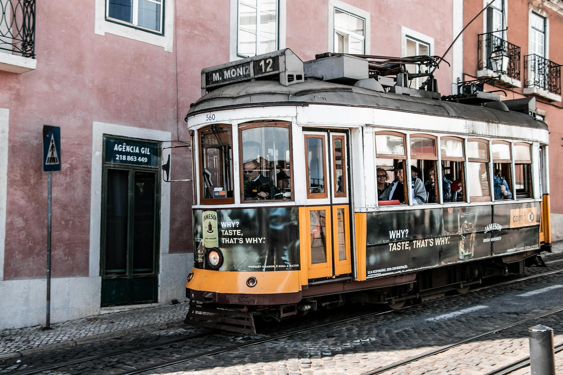 Elétrico Clássico Em Lisboa: Como Andar No Tram 28 Sem Ser Esmagado 2 Iconic vintage tram traversing a Lisbon street with cobblestone path.
