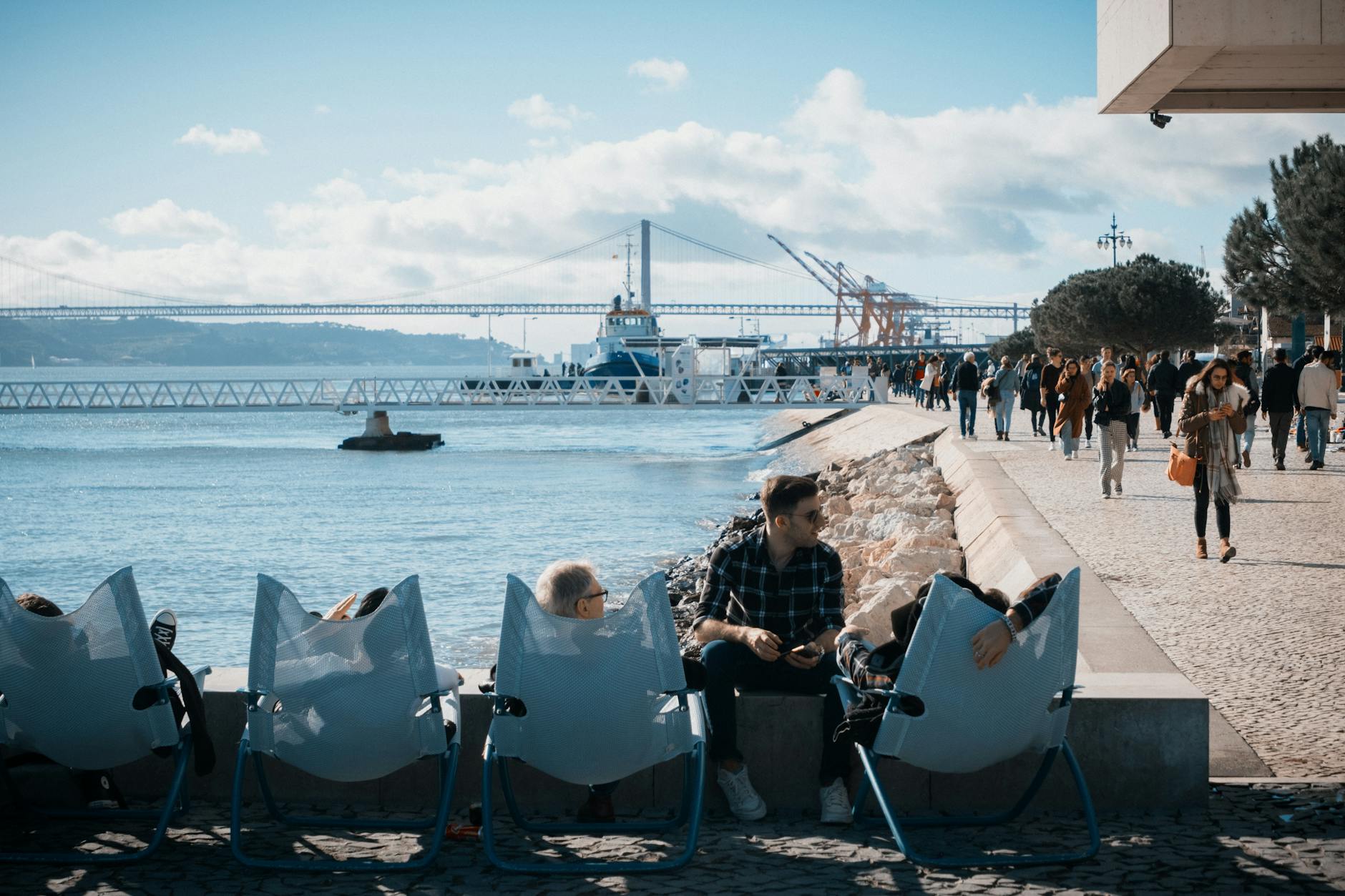 People relaxing on chairs by the Tejo River in Lisbon, with a busy waterfront and iconic bridge view.