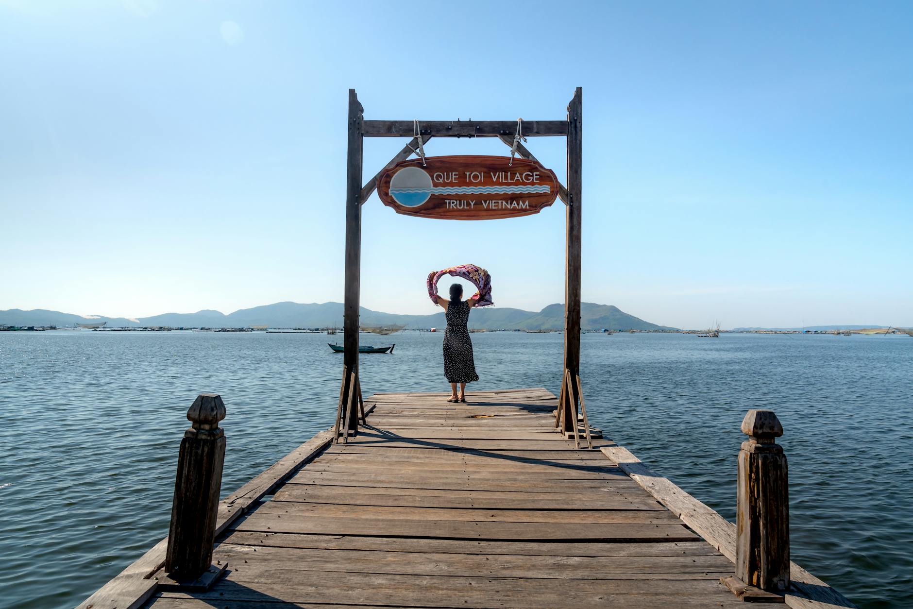 Scenic view of a woman standing on a dock at Que Toi Village by the ocean under a blue sky.
