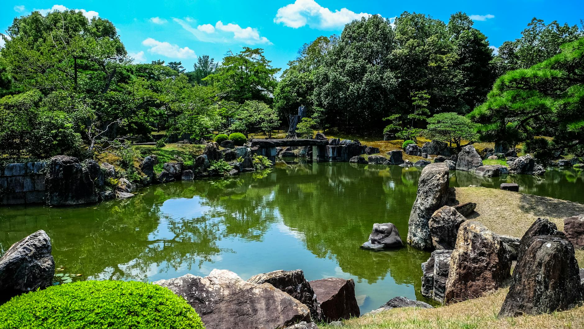 Os Melhores Jardins Em Lisboa Para Um “Reset” Mental 3 Tranquil Japanese garden with rocks, pond, and lush greenery under a clear sunny sky.