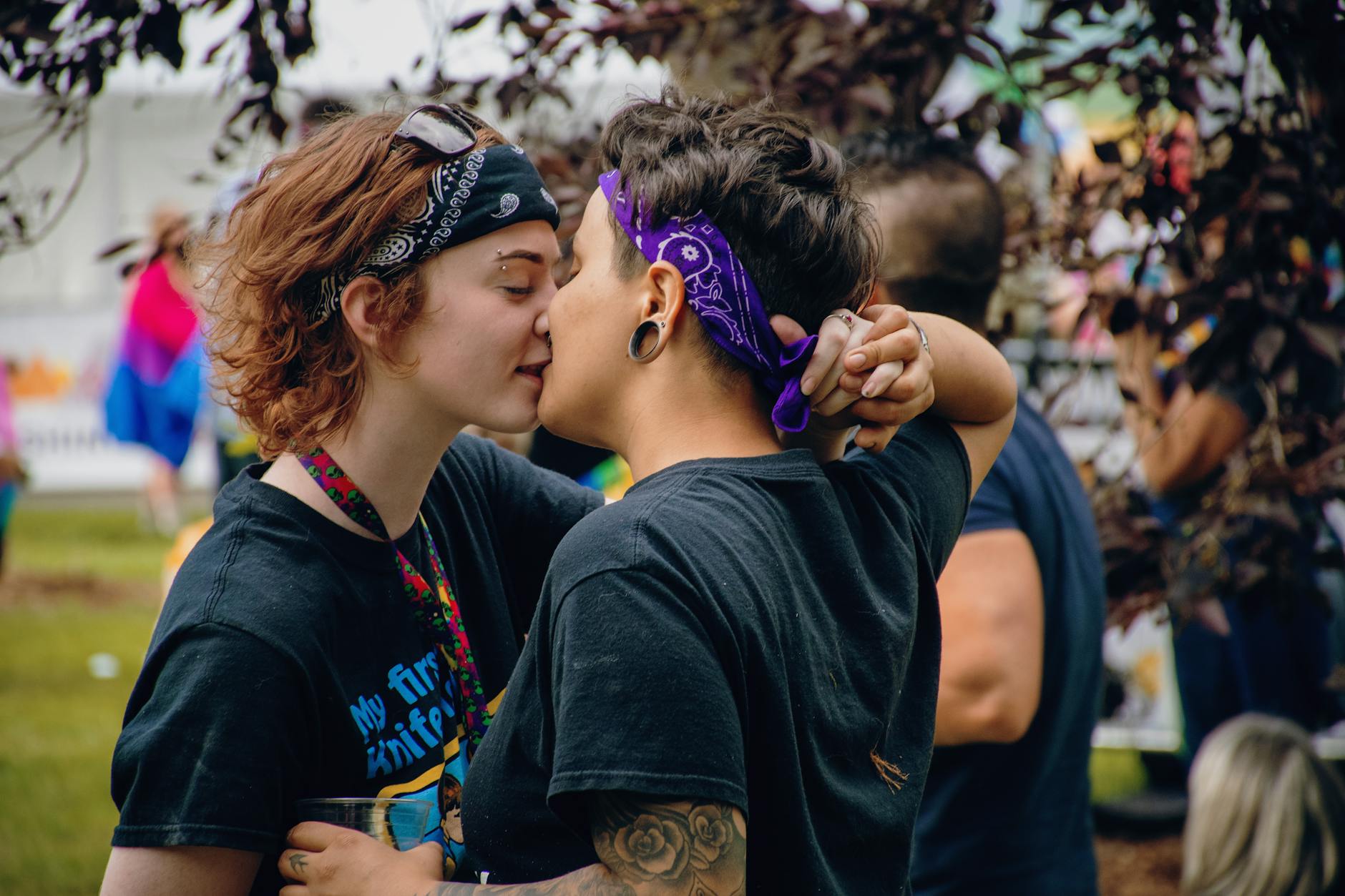 Two women share a kiss at a vibrant outdoor pride festival, celebrating love and inclusivity.