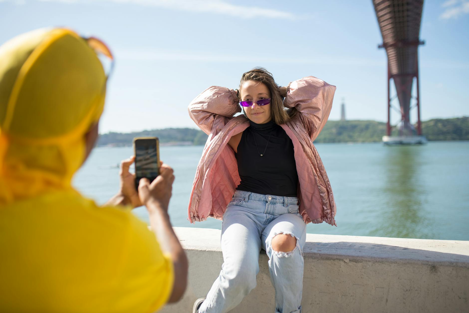 Woman in pink jacket and sunglasses posing near Lisbon's iconic bridge, urban style captured.