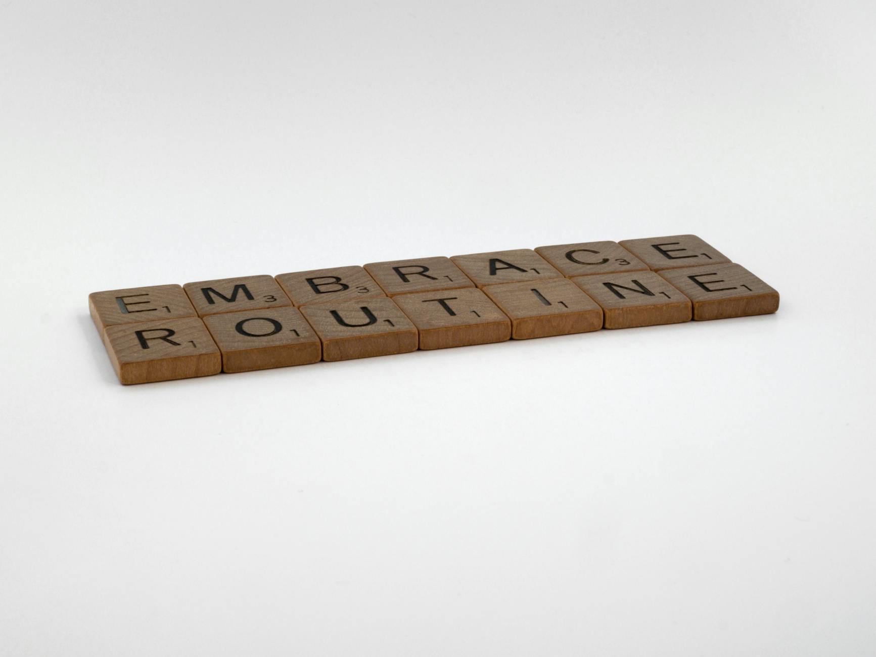 Wooden Scrabble tiles spelling 'Embrace Routine' on a minimalist white background, emphasizing typography.