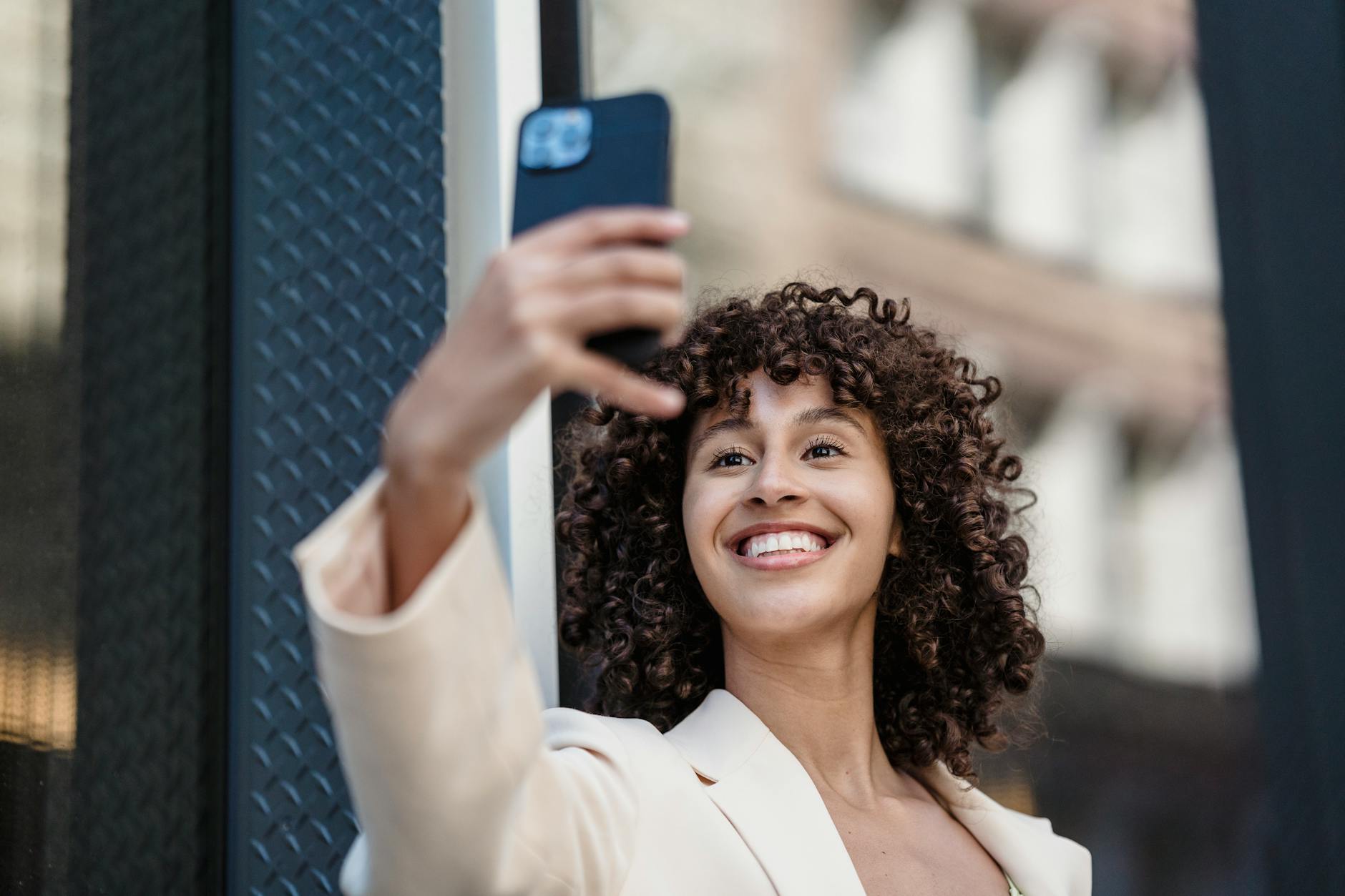 Young ethnic female with toothy smile and curly hair taking self portrait on mobile phone in city